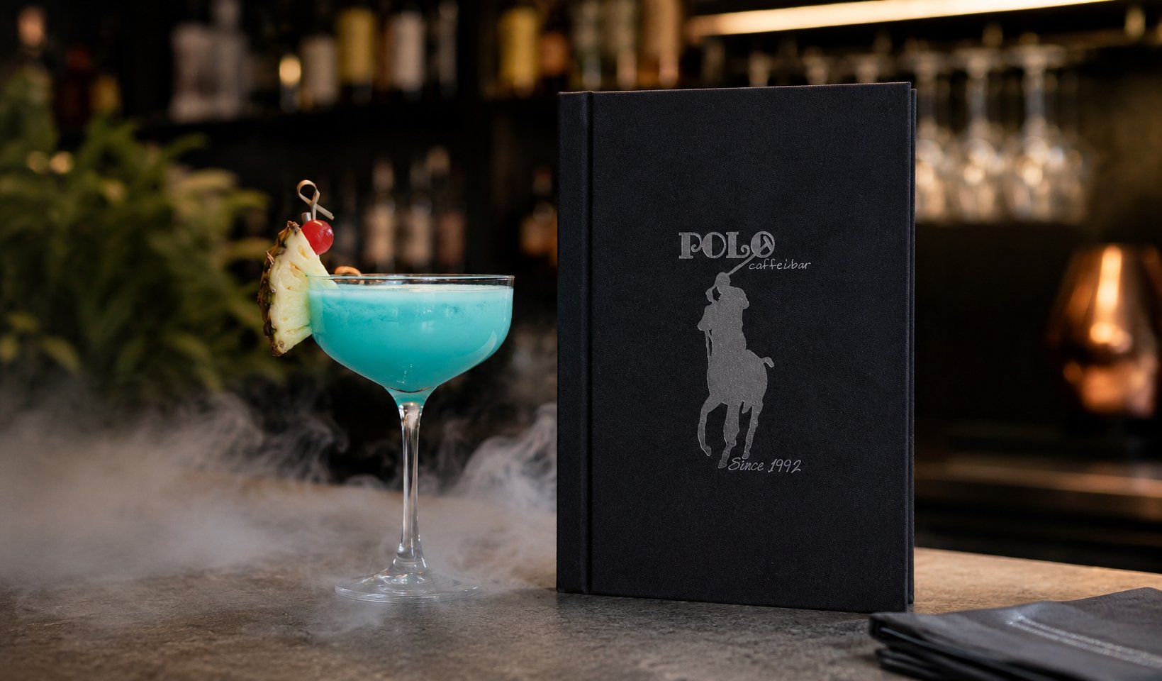 Črn usnjen meni z logotipom "Polo Caffe Bar" ob modrem koktejlu z dekoracijo in dimnim učinkom na pultu v elegantnem ambientu bara.
